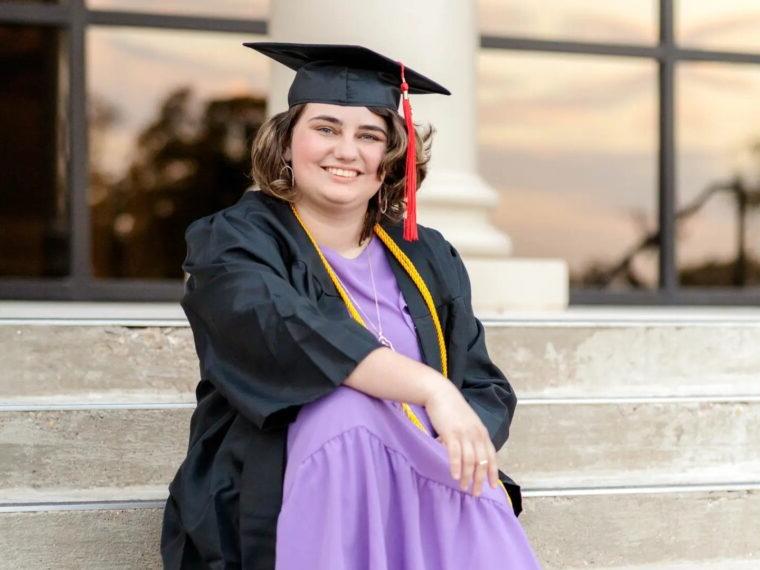 Madison Lewis, wearing her cap and gown, sits on the steps of the Skiles Building